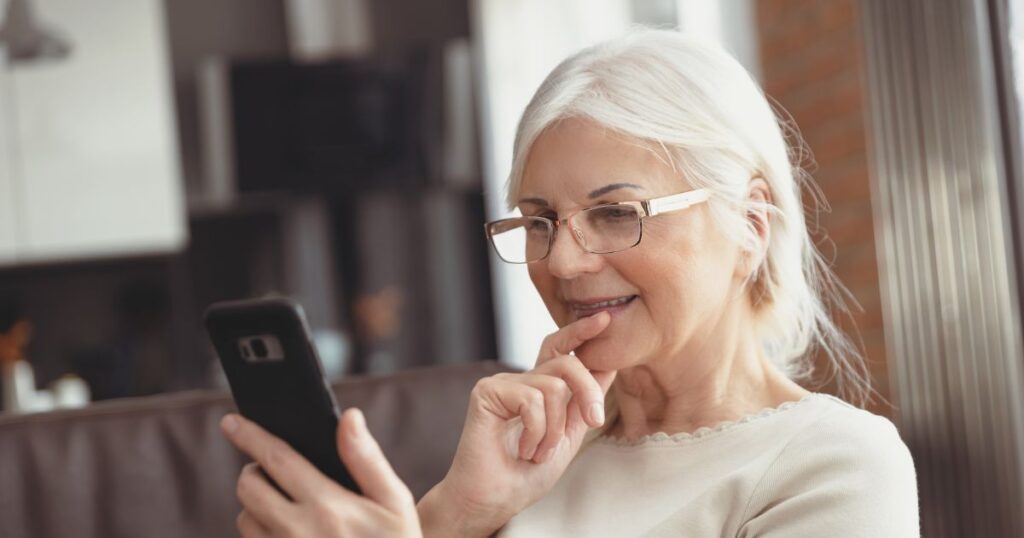 Ältere, weiblich gelesene Person, die lächelnd auf ein Smartphone schaut