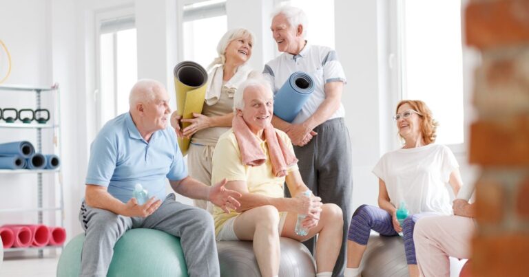 5 ältere Personen in Sportklamotten, teilweise auf Gymnastikbällen und Yogamatten in der Hand.