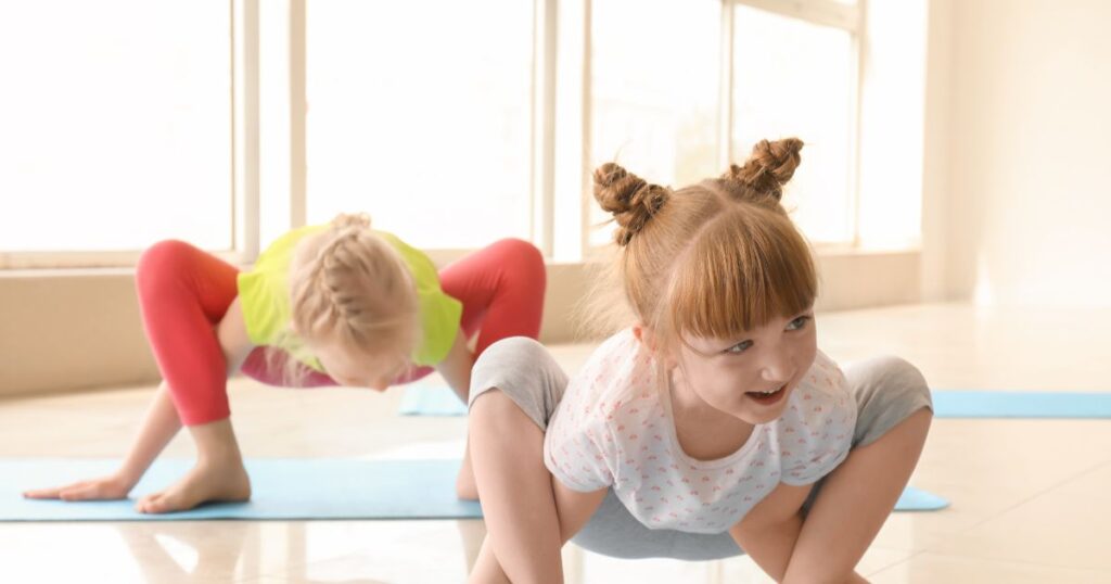 Zwei Kinder auf Sportmatten in einer Yogapose, wo die Arme durch die Beine in der Hocke geschoben