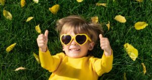 Kleinkind mit gelber Sonnenbrille und gelben Pullover liegt auf dem Rücken auf einer grünen Wiese, lacht und zeigt beide Daumen in die Höhe