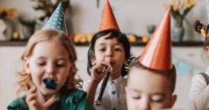 Drei Kinder, die einen Partyhut tragen und in eine Tröte pusten.