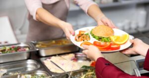Nahaufnahme eines Tellers mit einem Burger und Gemüse, der von einer Person an eine andere gereicht wird.