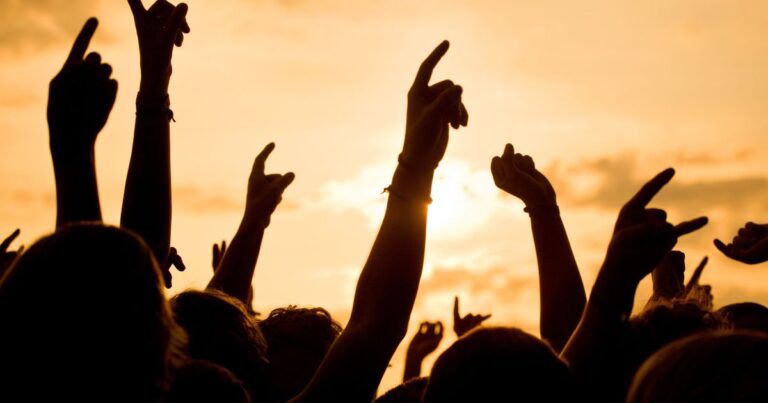 Im Hintergrund ein orange-gelber Himmel, im Vordergrund herausgestreckte Hände wie auf einem Konzert