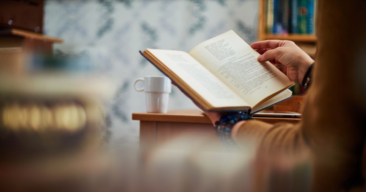 Person liest Buch, daneben steht ein Kaffee auf einem Tisch.