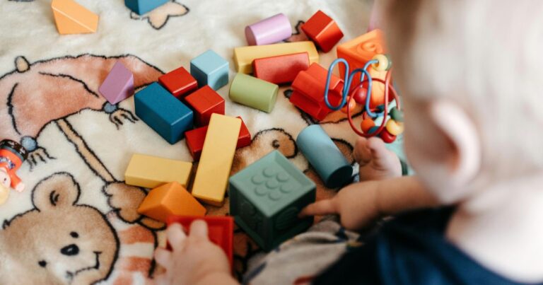 Bunte Bauklötze liegen auf einem Spielteppich. Kleinkind greif danach.