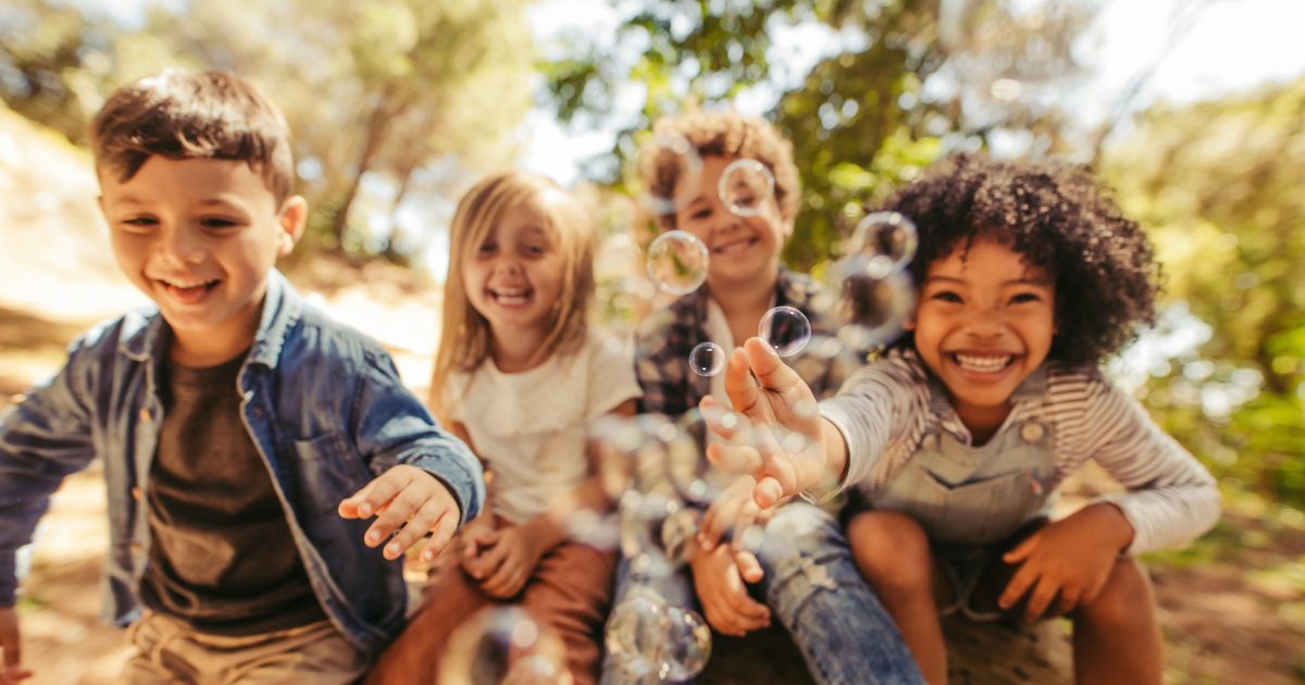 Vier kinder sitzen nebeneinander. Lachend, mit Seifenblasen im Hintergrund.