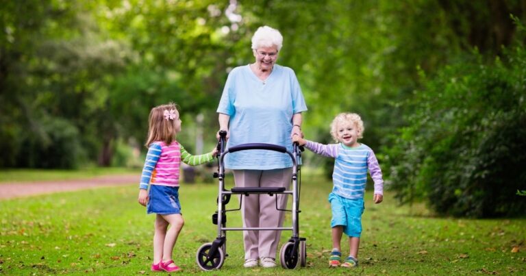 Seniorin schiebt Rollator draußen. An ihrer Seite 2 Kinder, die sie freudig anschauen.
